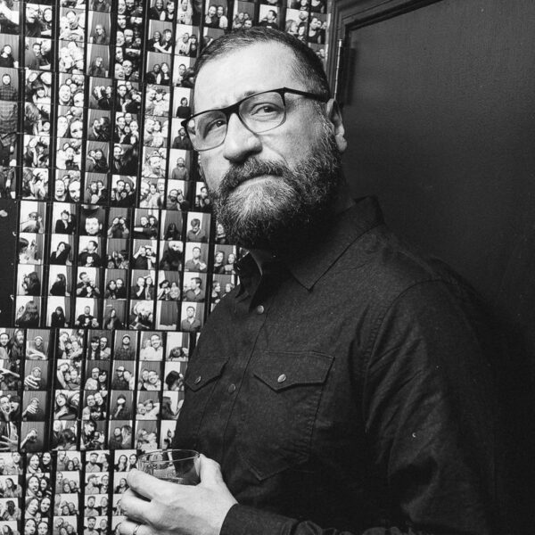 Black and white photo of an individual with a beard wearing glasses and a dark shirt, standing in front of a wall covered in numerous small photos. The person is holding a drink and looking off camera to their left.