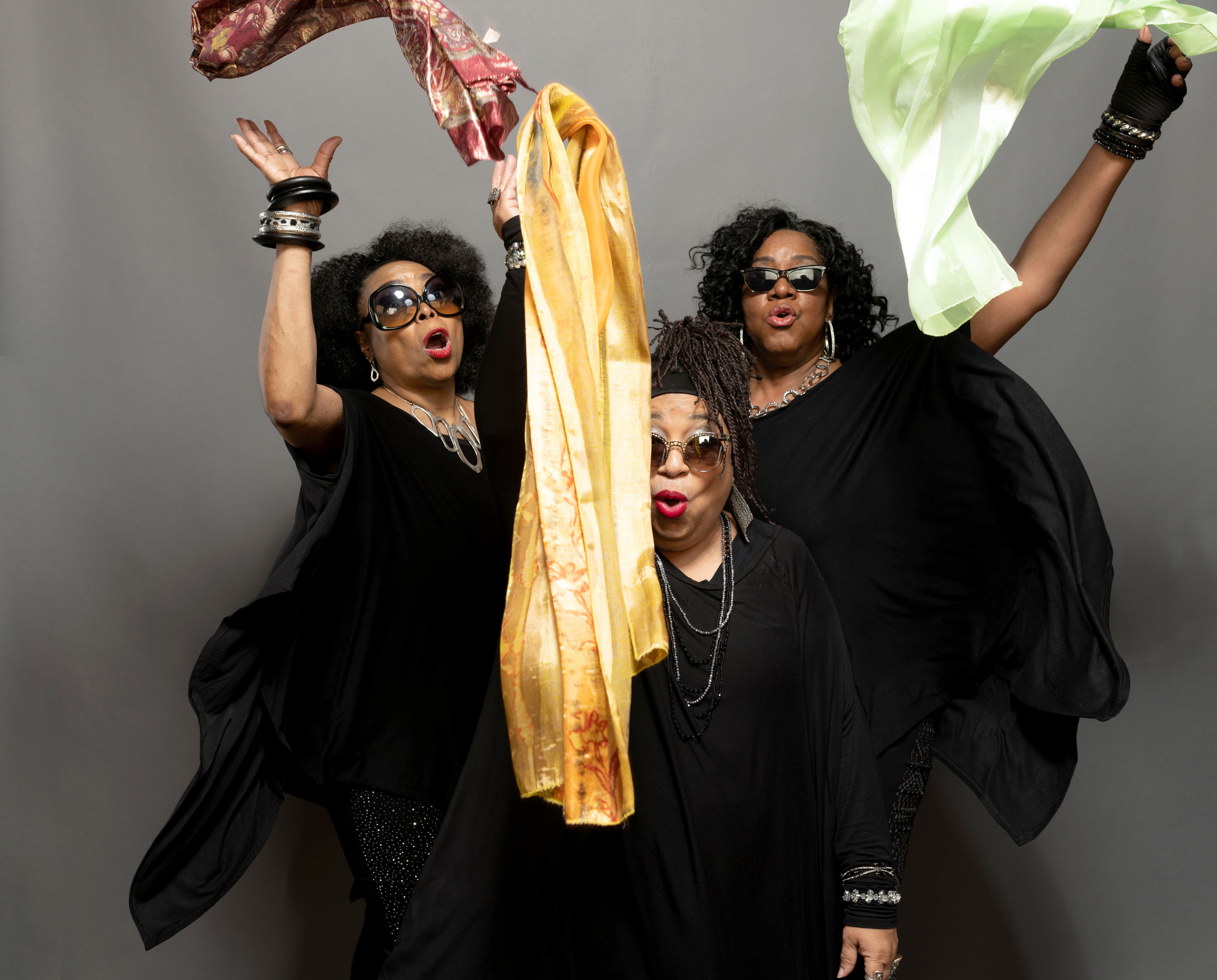 Group portrait of three brown-skinned female presenting people dressed all in black against a gray screen. Three scarves are captured mid-air