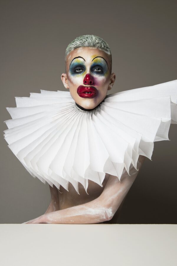 Three-quarter portrait of a person with short blond hair wearing clown makeup and an oversized ruffle collar