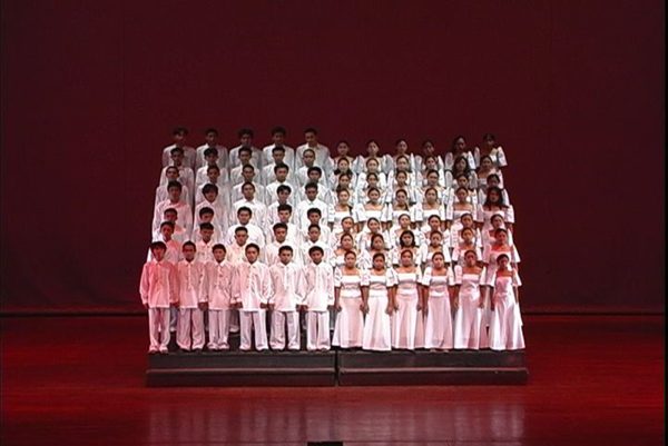 A choir dressed in white on elevated stands on a stage cast in red light