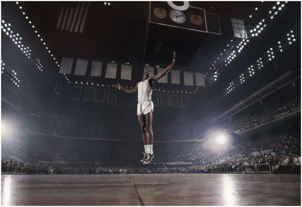 a basketball player hovering above our line of sight in a cruciform position in an arena filled with spectators