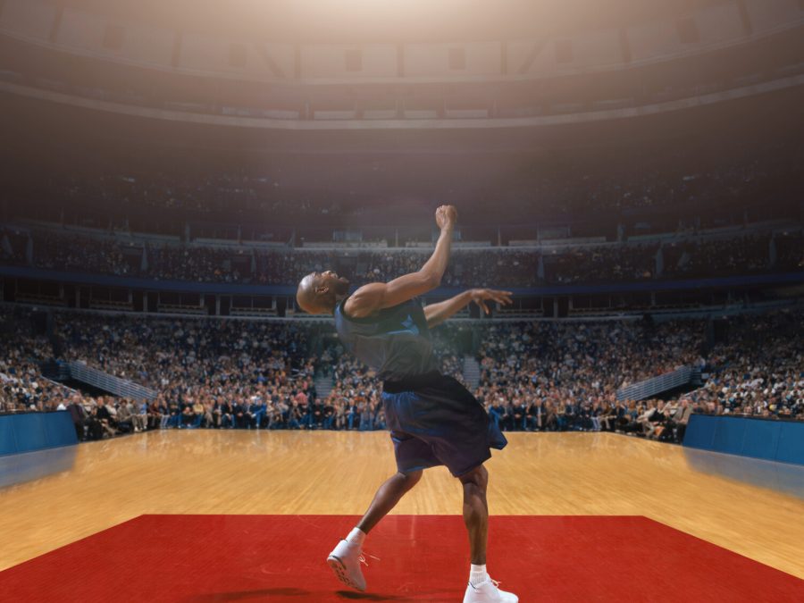 A Black basketball player on a court alone looks like he's about to do a backward flip in front of a crowded stadium.