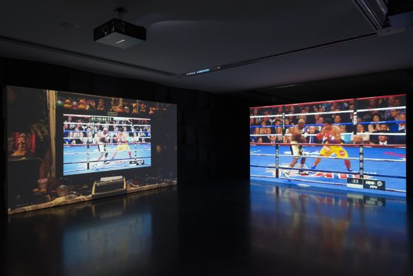 In a dark room two screens display a boxing match. The screen on the right displays it in full; on the left, it is displayed on a computer screen surrounded by a variety of objects, including rubber ducks.