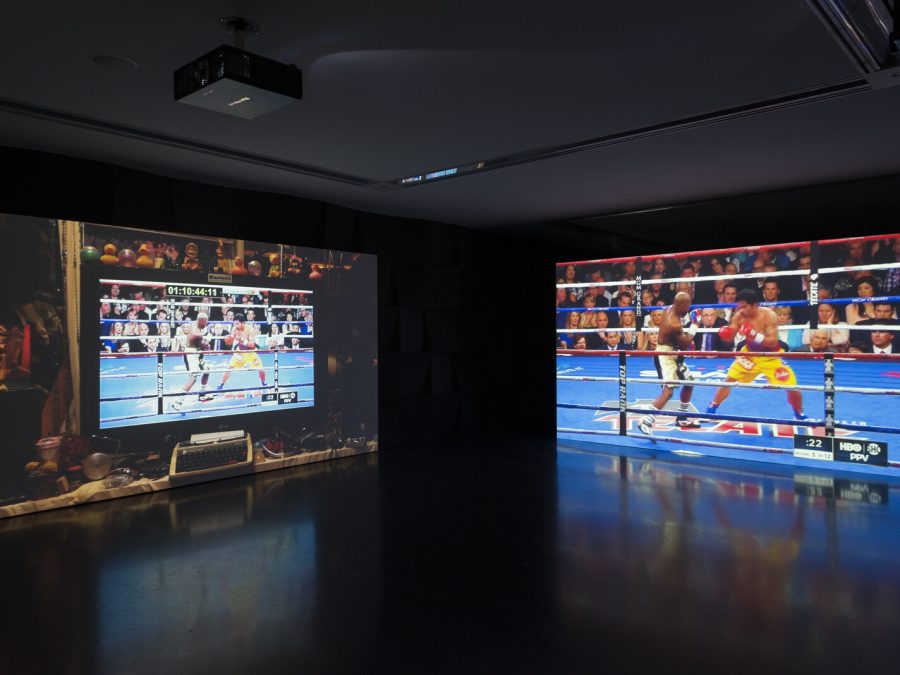 In a dark room two screens display a boxing match. The screen on the right displays it in full; on the left, it is displayed on a computer screen surrounded by a variety of objects, including rubber ducks.