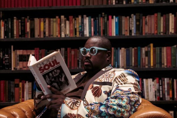 A brown-skinned male presenting person in a colorful suit sitting in a leather chair reading. A library of books appears behind them