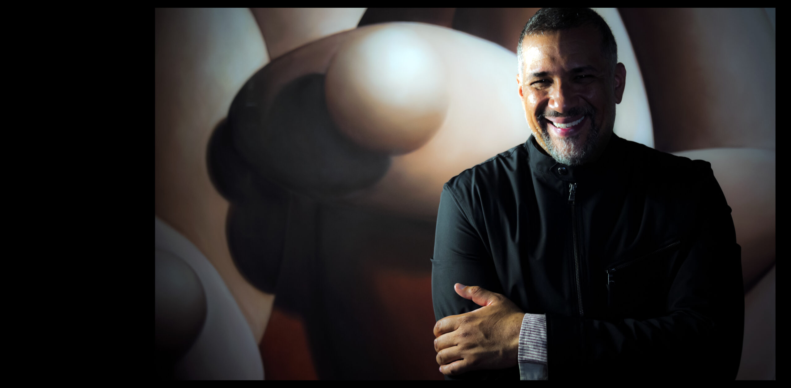 A male-presenting person with light-brown skin in front of an abstract mural