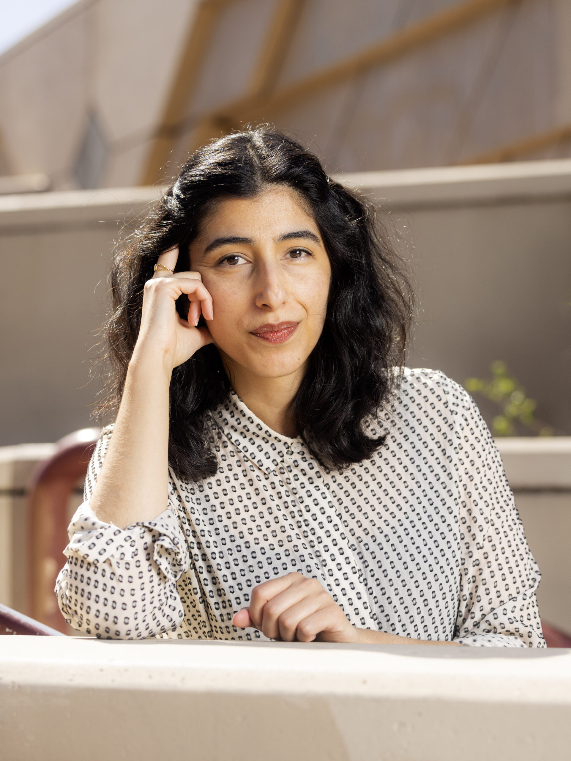 Portrait of a female-presenting person with olive skin and dark brown hair. Their right arm rests on a ledge with their hand to their face in a contemplative pose