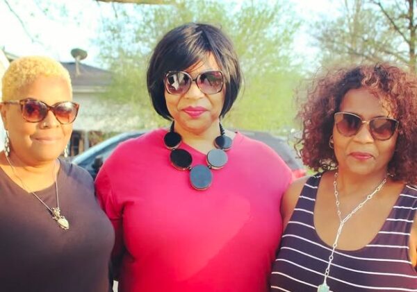 A group photo of three brown-skinned female-presenting people wearing sunglasses with their arms around each other