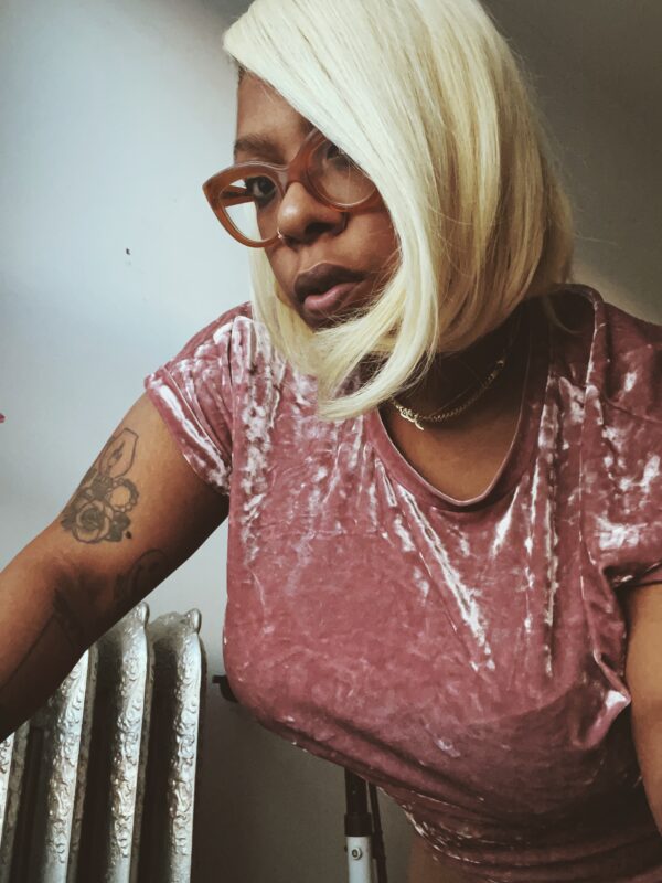 A female presenting person with medium-brown toned skin and platinum blonde hair leans in to the camera for a portrait.