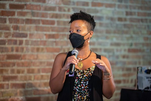 A person stands against a brick wall holding a microphone and wearing a black medical mask.