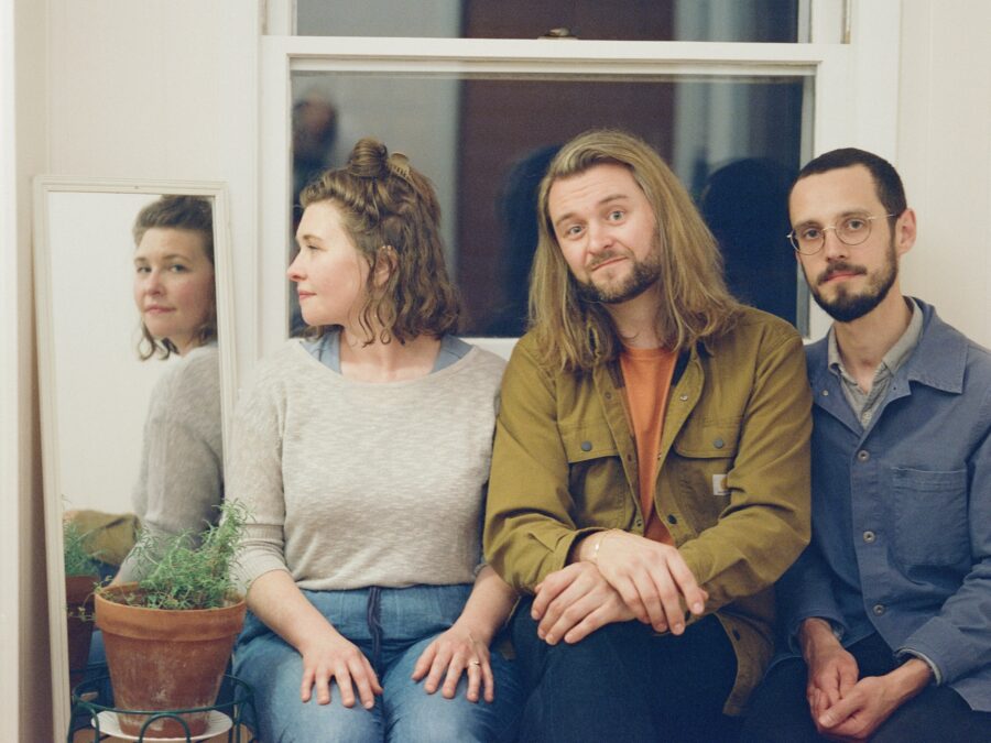 A portrait of three people sitting in front of a window. The sole female-presenting person turns their head to look in a mirror, which reflects their face towards the photographer