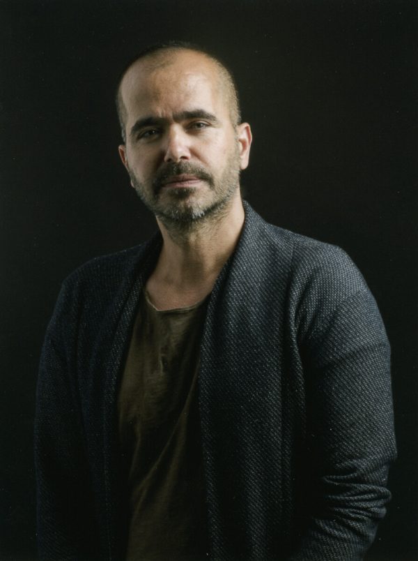 Moody portrait of a man with light brown skins and a shaved head wearing a dark shirt and cardigan against a black background