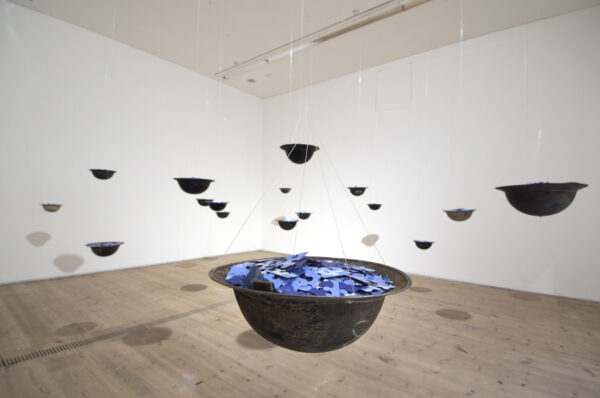 Installation of hanging upside down metal helmets filled with blue puzzle pieces in a white walled gallery space