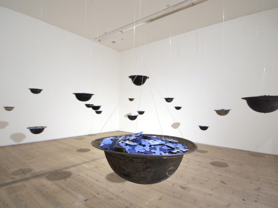 Installation of hanging upside down metal helmets filled with blue puzzle pieces in a white walled gallery space