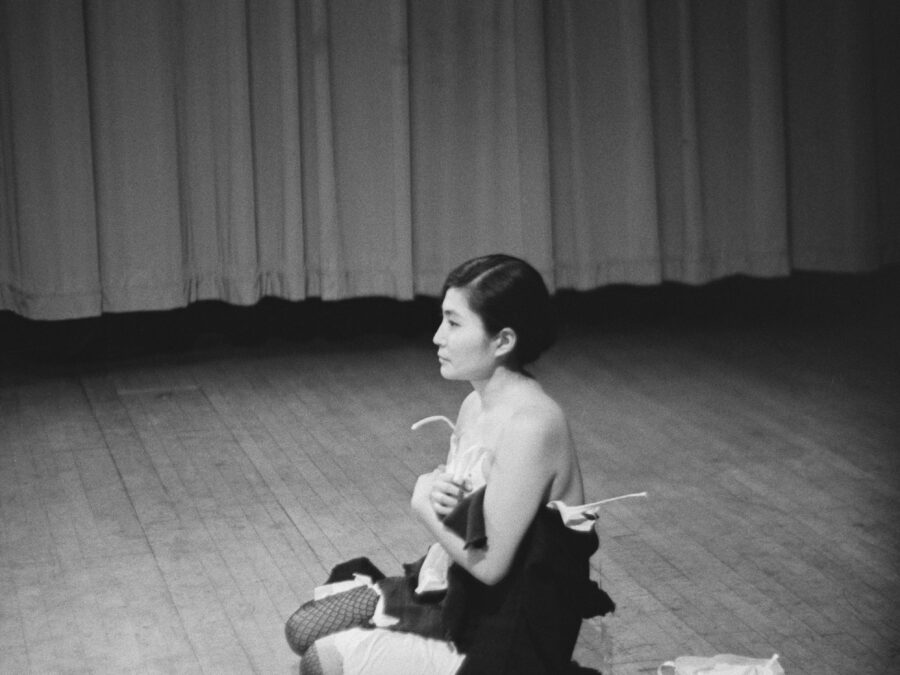 Archival photo of Yoko Ono sitting on the ground wearing scraps of clothing