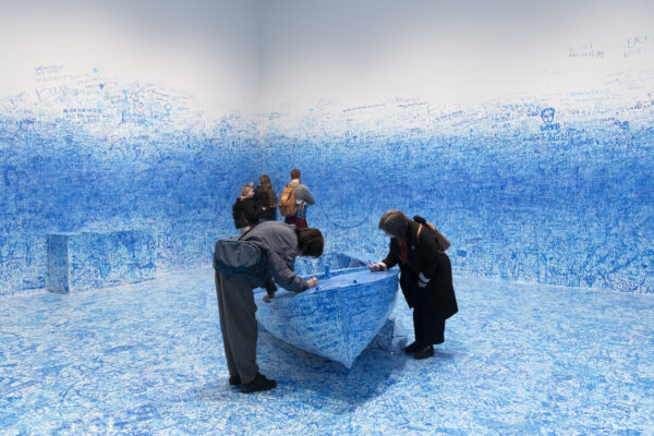 People write on a boat and the walls in a room filled with blue handwriting and doodles on the floor, boat, bench, and halfway up the otherwise white wall.