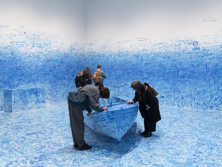 People write on a boat and the walls in a room filled with blue handwriting and doodles on the floor, boat, bench, and halfway up the otherwise white wall.