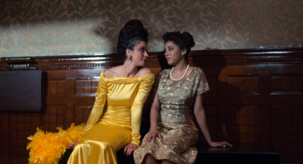 Two brown-skinned women sit on a wooden bench. One wears a bright yellow shiny evening dress with a feather boa next to her; the other wears a floral dress and a pearl necklace.