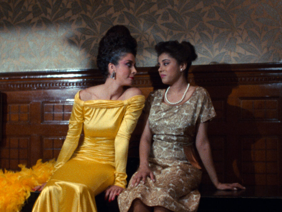 Two brown-skinned women sit on a wooden bench. One wears a bright yellow shiny evening dress with a feather boa next to her; the other wears a floral dress and a pearl necklace.