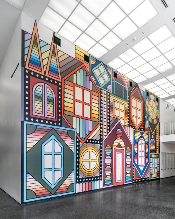 A two-story wall filled with a illustrated mural of colorful dormers, some upright, some upside down, featuring different style windows around a door in the middle.