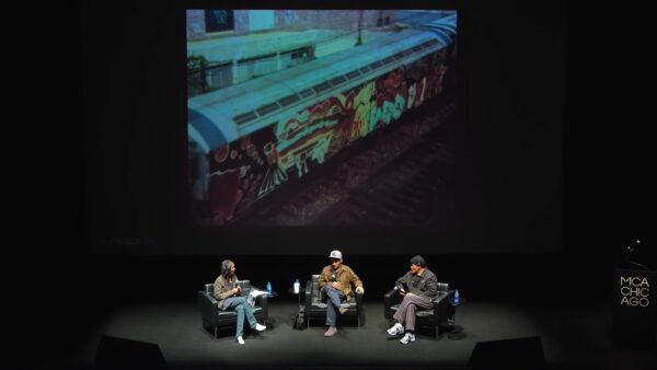 Three people sit on a stage an image of a train covered in graffiti is displayed on a large screen behind them
