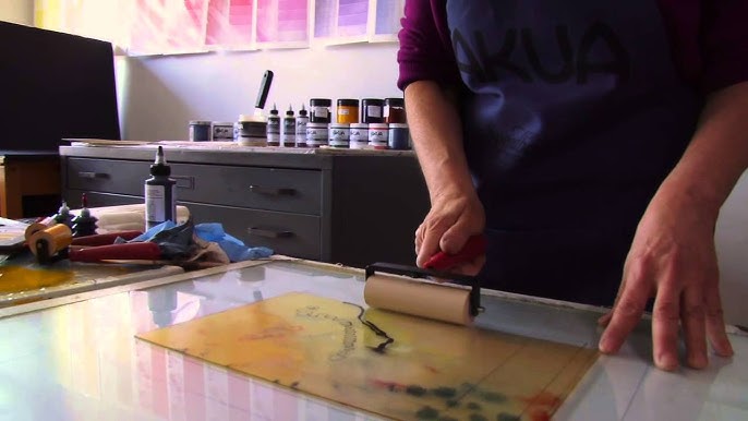 A hand moves a roller over a sheet of paper in a studio filled with art making supplies