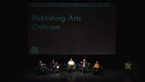 A group of people sit on a stage in a row. Behind them a screen casts a slide with the words Publishing Arts Criticism