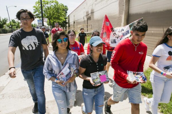 A group of teens holding postcards and a banner walk outside a brick building