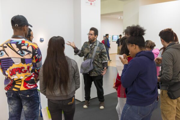 A group of teenagers gather around a person in a camo collared shirt in a gallery space