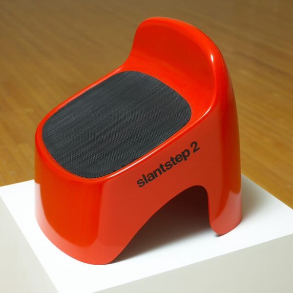 A red slanted stepstool or booster chair with a black rubber slip pad on the top on a white pedestal. On the side black text reading slant step 2 appears.