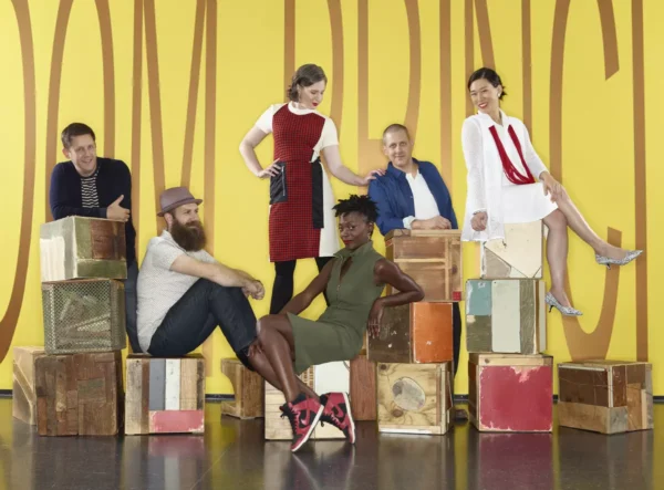 A group of seven people pose on and around boxes against a colorful yellow wall with large, indecipherable letters on it