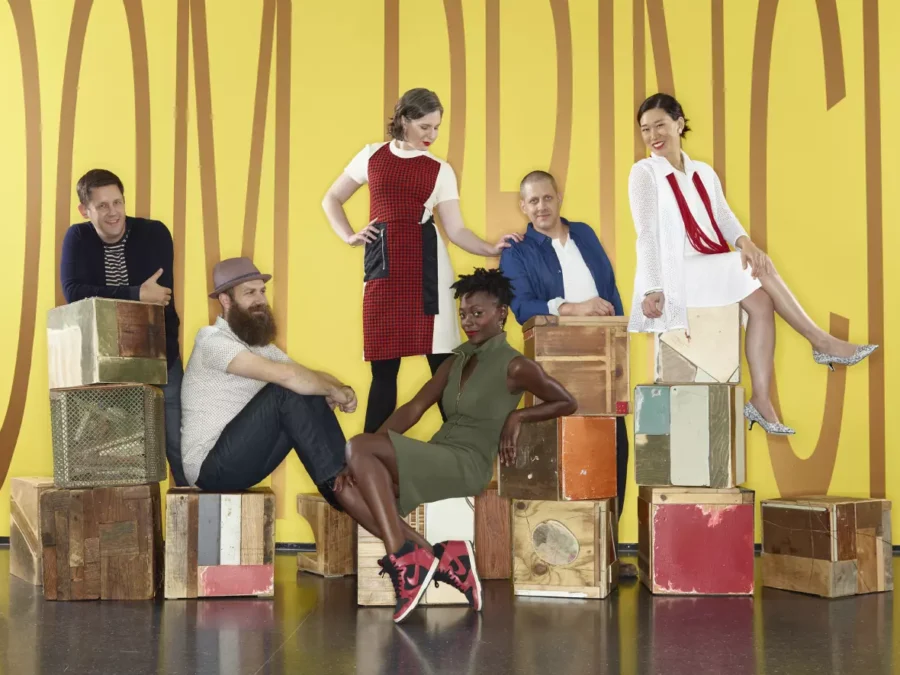 A group of seven people pose on and around boxes against a colorful yellow wall with large, indecipherable letters on it