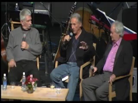 Three men with various tones of ivory skin and grey hair sit on a stage holding microphones