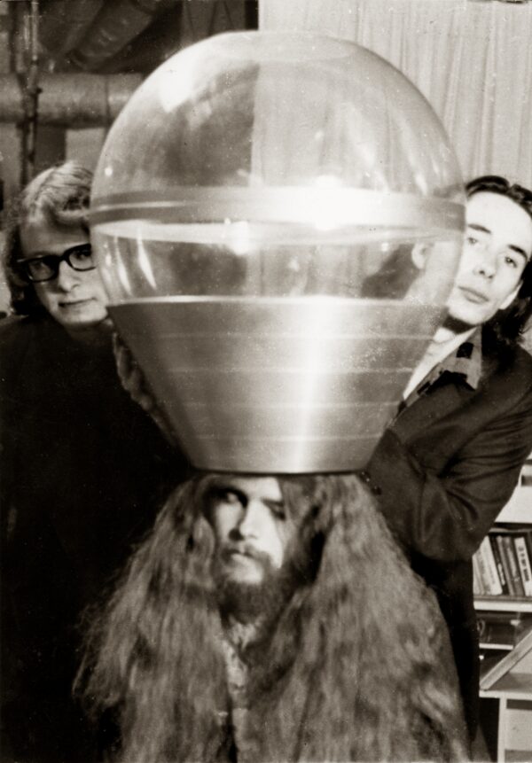 Black and white photo of three people posing underneath and on either side of a large clear sphere with an inverse metallic truncated cone attached at the bottom, resembling a large lightbulb.