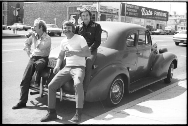 Black and white photo of three men with light skin sitting on an old Chevy