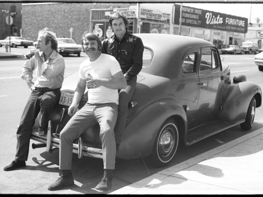 Black and white photo of three men with light skin sitting on an old Chevy