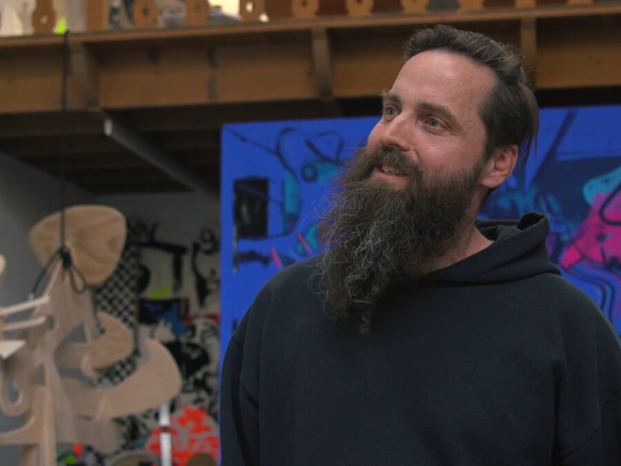 A male-presenting person with light skin and a large brown beard in a studio filled with sculptures and paintings