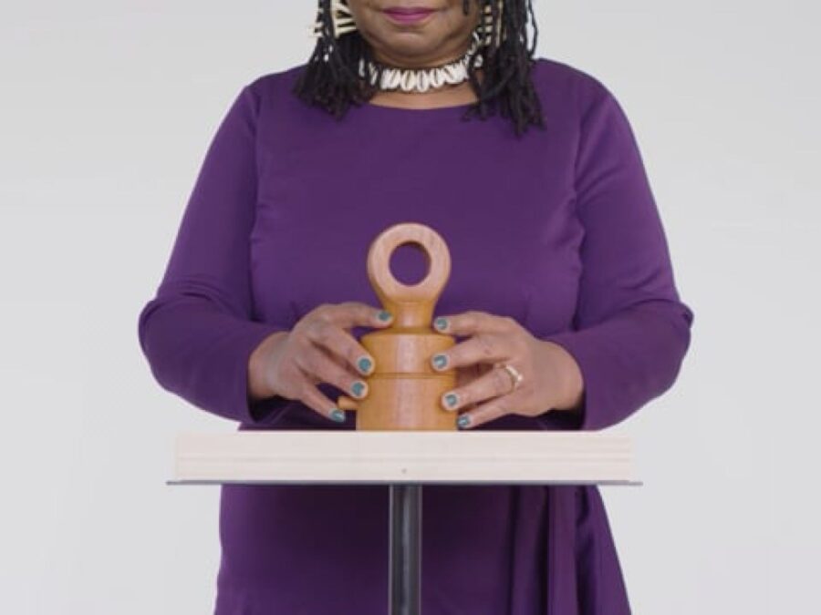 The torso of a person with light brown skin wearing a purple shirt and a puka shell necklace rests their hands on either side of a small clay sculpture with a looped top handle.