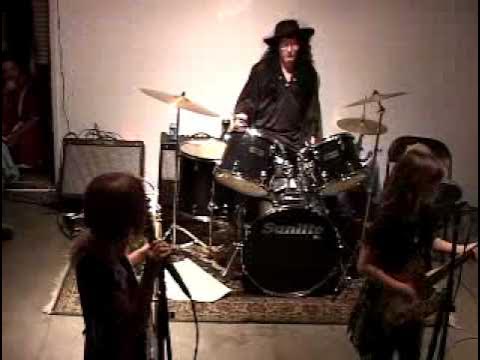 Grainy video still of a live concert featuring a person dressed in all black and wearing a flat-brimmed fedora on the drums