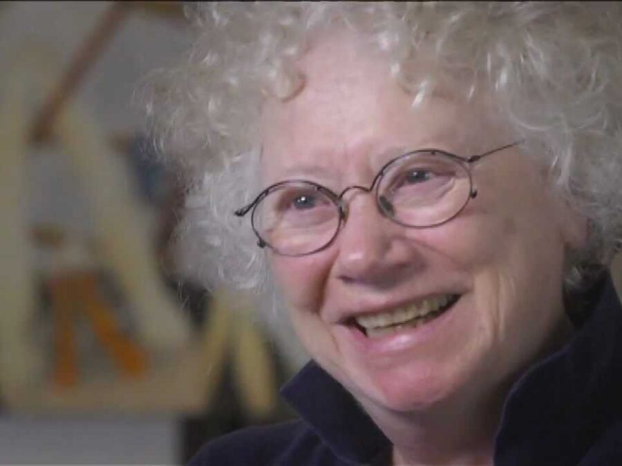 A woman with pale ivory skin and short curly white hair smiles toward something off camera