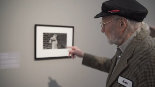 A man with grey hair wearing a suitcoat and hat with a red lightning bolt pin on it points at a photograph of a child in a grey room