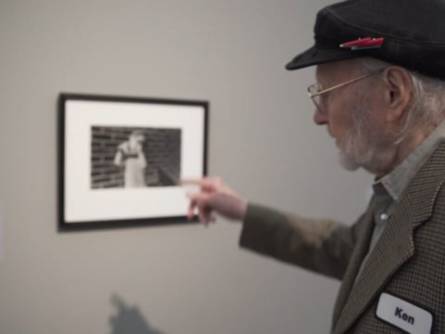 A man with grey hair wearing a suitcoat and hat with a red lightning bolt pin on it points at a photograph of a child in a grey room