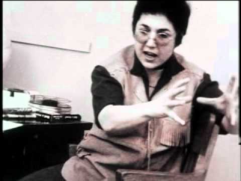 An archival still of Judy Chicago gesturing next to a desk