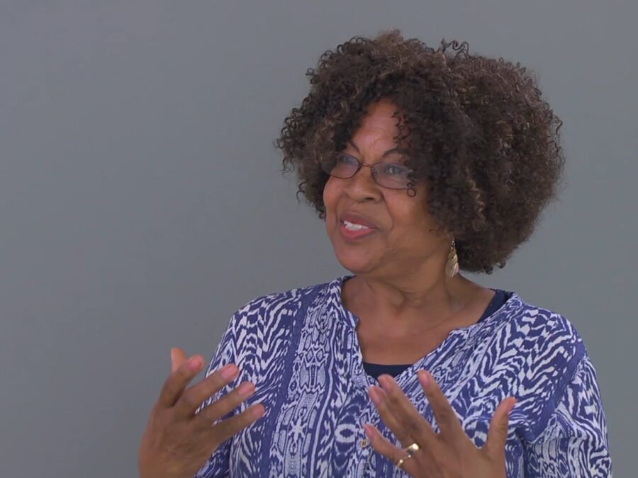 A female-presenting person with light-brown skin and a full head of short, curly hair against a grey screen gestures off camera to their right.