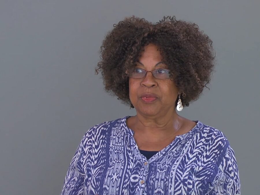 A female-presenting person with light-brown skin and a full head of short, curly hair against a grey screen looks off camera to their right.