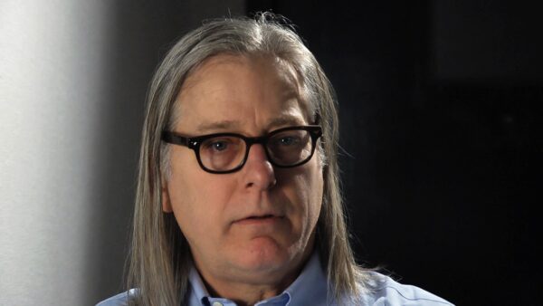 Closeup of a masculine person with glasses and long grey hair in a darkened room