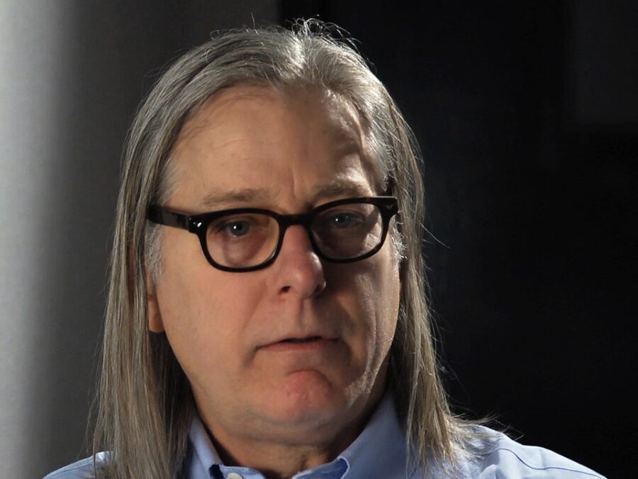 Closeup of a masculine person with glasses and long grey hair in a darkened room