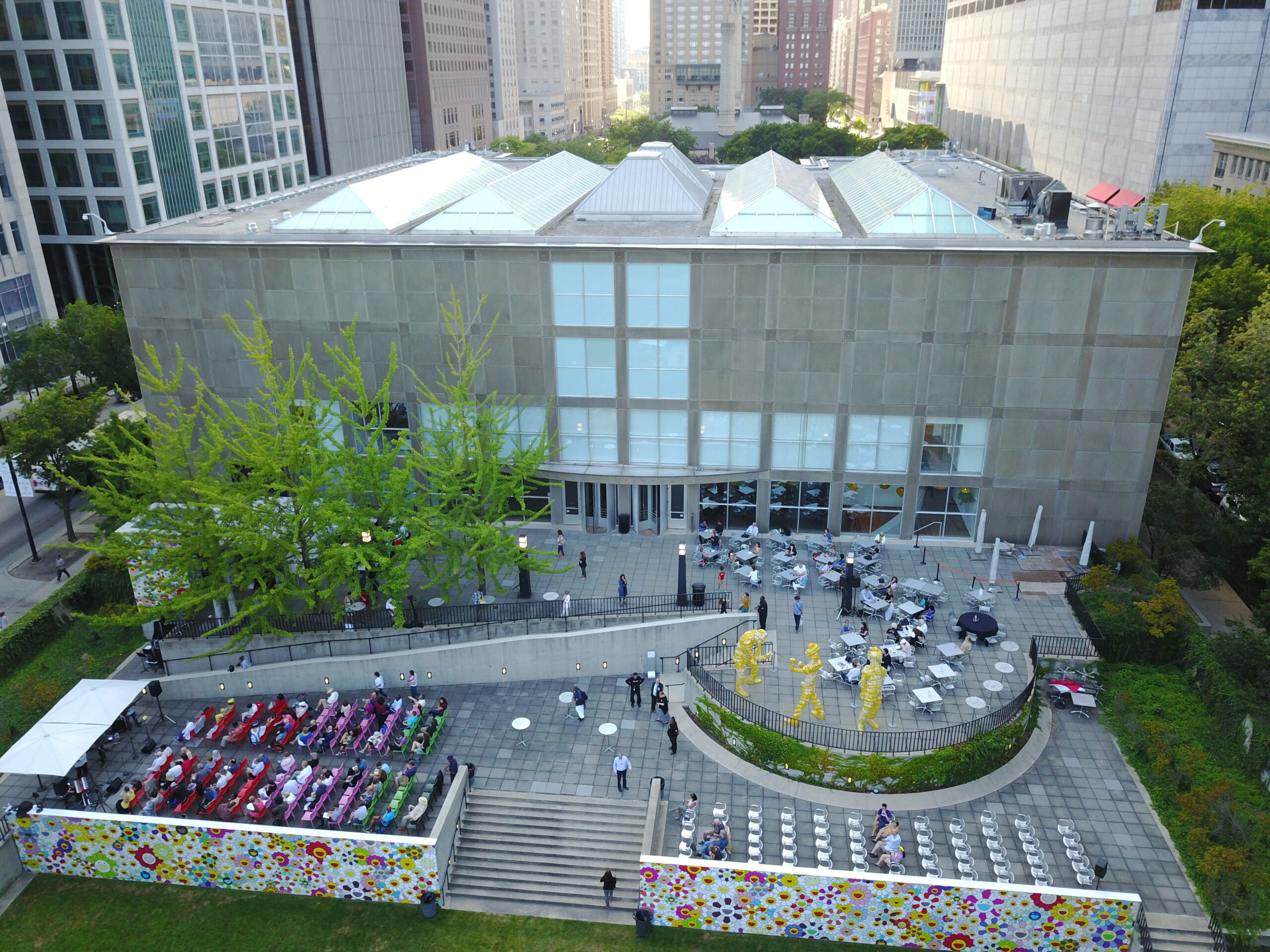 Aerial view of the back of a gray rectangular building, with an outdoor patio filled with tables and rows of chairs filled with a small grouping of people as well as steps down to a grassy garden that is out of view. Trees and grass are green.