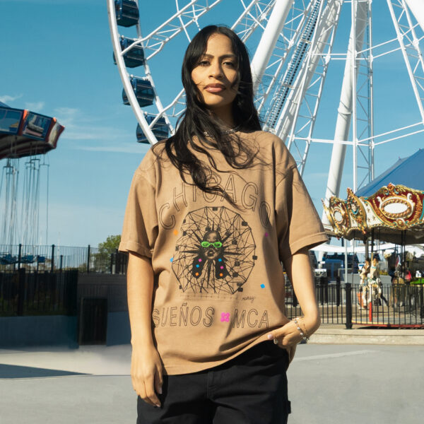 A person with long dark brown hair poses in a tan t-shirt in front of traditional fair rides like a ferris wheel and a carousel