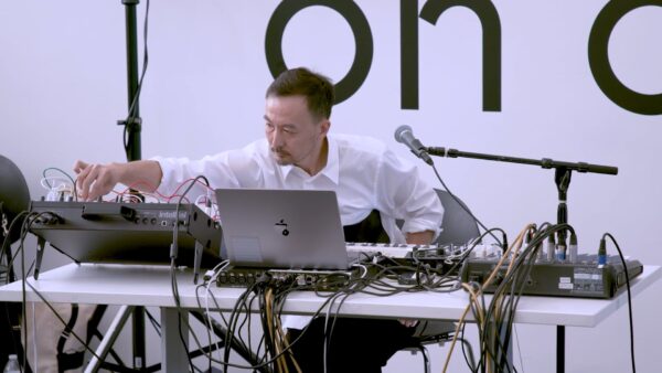 A masculine person sits at a table filled with recording and electronic equipment